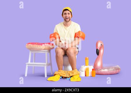 Male vacationer with beach accessories sitting on toilet bowl against lilac background Stock Photo