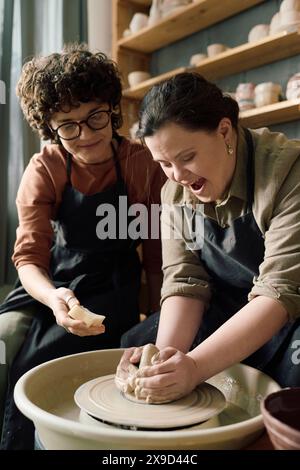 Young woman with Down syndrome sitting next to artisan enjoying pottery class in modern workshop Stock Photo