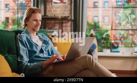 Young Handsome Adult Man Sitting on a Couch in Loft Living Room, Working from Home on Laptop Computer. Creative Male Checking Social Media, Browsing Internet. Urban City View from Big Window. Stock Photo