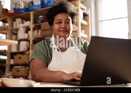 Warm toned portrait of African American senior woman using laptop in art studio lit by sunlight copy space Stock Photo