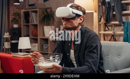 Television Sitcom Concept: Guy Using Virtual Reality Headset Eating Breakfast in Living Room And Smiling. Funny Sketch About Internet Addiction. Comedy Series on Network Channel, Streaming Service. Stock Photo