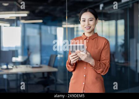 Portrait of a successful Asian female programmer with a tablet computer in her hands, an employee of a development company is smiling and looking at the camera near the window. Stock Photo