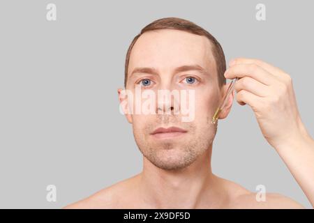 Man skin care routine. Oil pipette near male face. Hair serum product. Haircare facial beard treatment. Applying healthy cosmetics. Beauty skincare hydration. Isolated on grey background Stock Photo