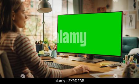 Over the Shoulder View of Creative Young Woman Sitting at Her Desk, Using Desktop Computer with Mock-up Green Screen. Female Caucasian Specialist Working on Computer with Chroma Key Display at Home. Stock Photo