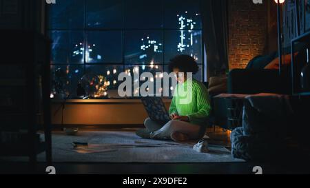 Young Black Woman Using Laptop Computer in Stylish Loft Apartment in the Evening. Creative Female Smiling, Checking Social Media, Browsing Internet. Night View From the Window. Stock Photo