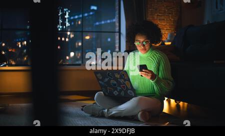 Black Female IT Specialist Coding on her Laptop in the Evening and Checking Notifications on Smartphone. Diverse Woman has Deadline Late at Night. Millennial People Working Concept. Stock Photo