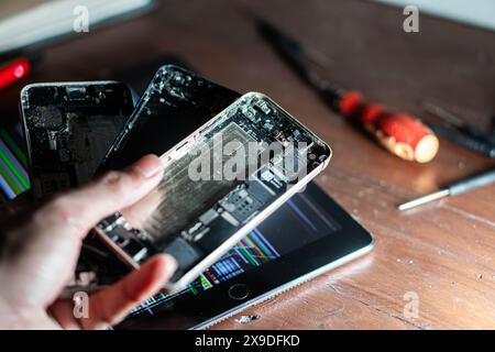 A person holding disassembled smartphone parts over a tablet with coding on the screen. Tools and electronic components are scattered on the wooden ta Stock Photo