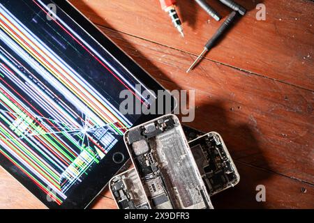A broken tablet with a colorful, distorted screen and two disassembled smartphones on a wooden surface, accompanied by repair tools. Stock Photo