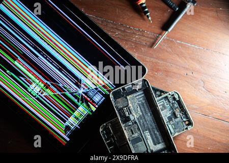 A broken tablet with a colorful, glitchy screen next to disassembled smartphones on a wooden surface. Tools for repair are visible in the background. Stock Photo