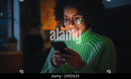 Cinematic Portrait of Black Female with Curly Hair Sitting on the Floor at Home and Using Smartphone. Diverse Woman Receives Good News and Chatting with Friends Via Social Media. Stock Photo