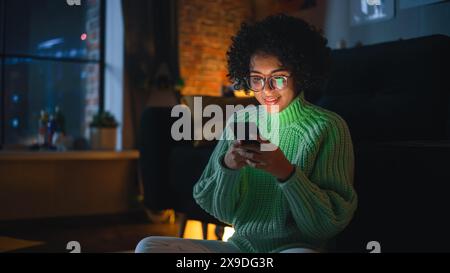 Latin Concentrated Woman Typing Message on her Smartphone in the Evening at Home. Internet Addicted Female Using Mobile Phone App for Online Shopping, Social Media, Browsing. Stock Photo