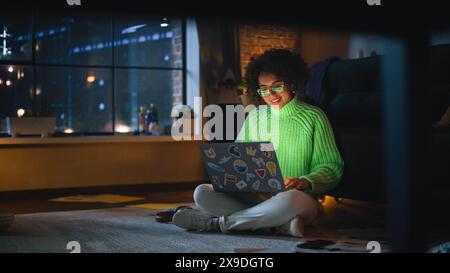 Positive Latina Female Using Laptop with Stickers at Home. Woman Resting on the Floor and Messaging on Computer with Smile on Face. Evening City View Behind the Big Window. Stock Photo