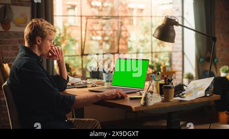 Millennial Designer Office: Caucasian Male Artist Sitting at His Desk Working on Laptop Computer With Green Screen Chroma Key on Display. E-Commerce and Smart People with Fresh Ideas Concept. Stock Photo
