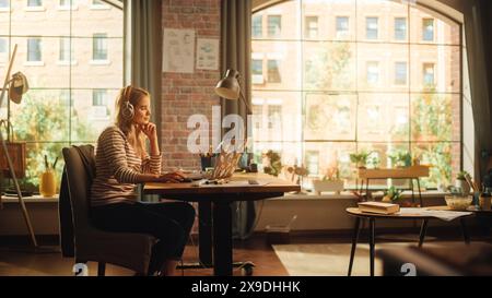 Young Pretty Woman with Blond Hair Wearing Headphones and Working from Home on Laptop Computer in Stylish Loft Apartment. Creative Female Smiling, Checking Social Media, Browsing Internet. Stock Photo