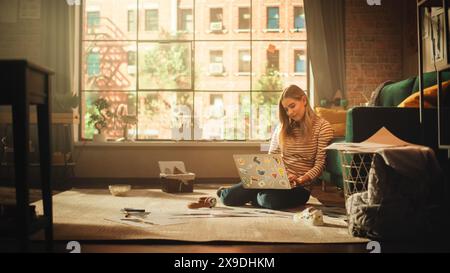 Beautiful Woman Using Laptop at Home and Working Remotely. Young Adult Woman Smiling and Sitting on the Floor Using Internet, Writing Report, Finishing Project. Good Sunny Day for Productive Work. Stock Photo