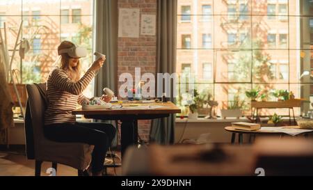 Woman Sitting Behind the Desk at Home or Creative Agency, Using Virtual Reality Headset and Controllers. Young Female Working from Living Room, Experiencing Futuristic Social Media Services. Stock Photo