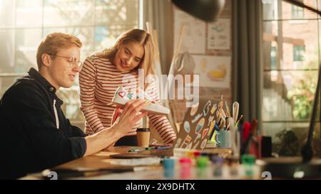 Young and Positive Female Artist Looks at Screen and Shares Ideas With her Male Colleague Who Uses Laptop Computer. Designers Creating New Model of Shoes. Modern Professionals Concept. Stock Photo