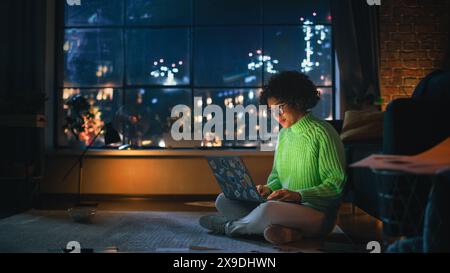 Black Female Student in Cozy Green Jumper Working On Her Laptop In the Evening. Smart Woman Thinking, Smiling, Answering Emails. Girl Study Late at Night in Loft Apartment Living Room. Stock Photo