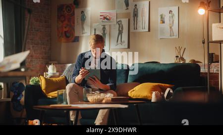Caucasian Male Designer Draws on Digital Tablet, while Sitting on Sofa in Relaxed Position. Loft Apartment Stylish Living Room. Smart People and New Generation Professionals Concept. Stock Photo