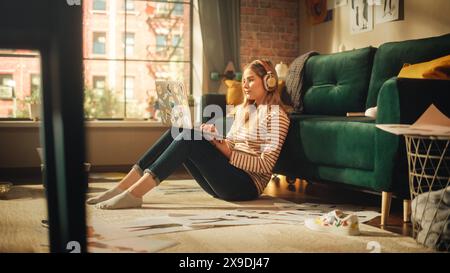 Blonde Attractive Woman Using Laptop Computer, Sitting on Living Room Floor in Cozy Stylish Loft Apartment. Beautiful Girl in Headphones is Thinking and Coding New Mobile App. Stock Photo