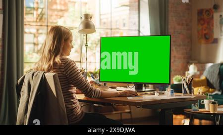 Over the Shoulder View: Creative Young Woman Sitting at Her Desk, Using Desktop Computer with Mock-up Green Screen. Female Caucasian Specialist Working on Computer with Chroma Key Display at Home. Stock Photo