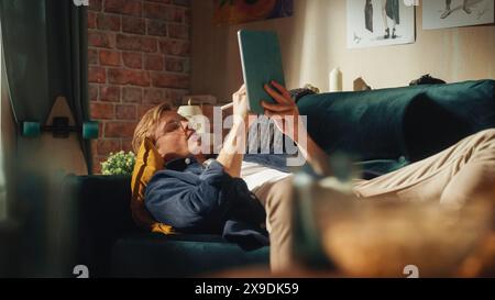 Focused Male Designer Works on Digital Tablet, while Resting on the Couch in Creative Office Environment. New Younger Generation Person Drawing, Writing, Creating New Things Concept. Stock Photo