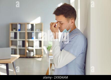 Stressed tired male doctor wearing blue uniform standing in hospital suffering from headache. Stock Photo