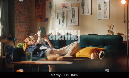 Caucasian Male Designer Draws on Digital Tablet, while Laying on Sofa in Relaxed Position. Loft Apartment Stylish Living Room. Smart People and New Generation Professionals Concept. Stock Photo