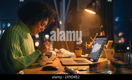 Side View of Active Female Designer Sitting in the Evening in Loft Studio and Working on Shoes Customisation in Front of Her Laptop. Next Generation and Trendy Design Concept. Stock Photo