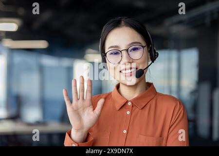 Webcam view, Asian woman with headset phone smiling and looking at camera waving hand, using video calling for customer consultation, customer support remotely. Stock Photo
