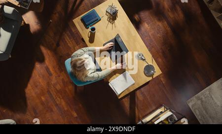 Top View of Home Office: Freelance Entrepreneur Sitting at Desk Works on a Laptop. Female Professional Works from Home, Manages Company Remotely, Designs Software. Remote Access Work Concept. Stock Photo