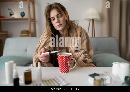tired sick woman sitting on sofa measures her temperature and drinks tea with flu lemon. Cold prevention Stock Photo