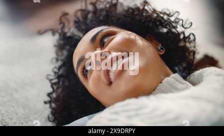 Close Up Portrait of Black Curly Female Talking with Somebody while Relaxing on the Floor at Home. Woman Plays with her Hair and Smiles Flirtaciously. Feeling of Falling in Love Concept. Stock Photo
