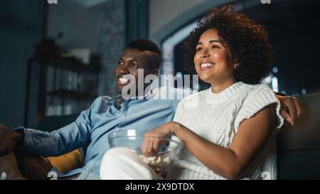 Black Couple Watching Comedy Movie on TV, Eating Popcorn while Sitting on Couch in the Apartment Late at Night. Laughing Boyfriend and Girlfriend Enjoying Funny TV Series at Home. Low Angle Shot. Stock Photo
