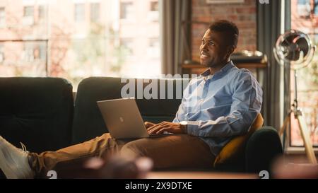 Handsome African American Man Working on Laptop Computer While Sitting on a Sofa in Cozy Living Room. Freelancer Working From Home. Browsing Internet, Using Social Networks. Student Studying Online. Stock Photo