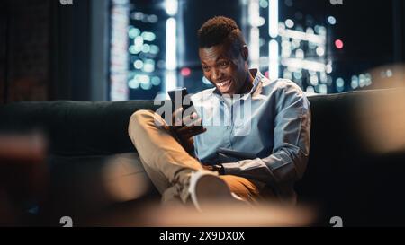 Excited Black African American Man Using Smartphone While Sitting on a Sofa in Living Room. Happy Man Smiling at Home and Chatting With Friends Over the Internet. Using Social Networks Concept. Stock Photo
