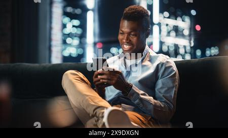 Excited Black African American Man Using Smartphone While Sitting on a Sofa in Living Room. Happy Man Smiling at Home and Chatting With Friends Over the Internet. Using Social Networks. Stock Photo