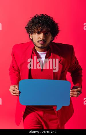 Handsome young Indian man in vibrant red suit holds a speech bubble against a vivid backdrop. Stock Photo