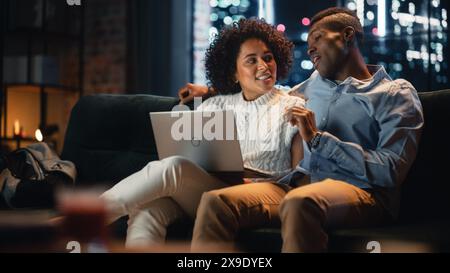 Black Couple in Love Use Laptop Computer, Have Fun, Laughs While Sitting on the Couch in the Cozy Loft Apartment. Couple Shopping on Internet, Using Social Media, Discussing Something. Stock Photo