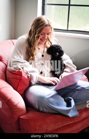 Woman sitting in a pink chair, using a laptop with a black dog o Stock Photo