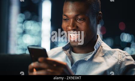 Close Up Portrait of Handsome Black Man Laughing While Using Smartphone in Cozy Living Room at Home. He's Having Fun while Browsing the Internet and Checking Videos on Social Networks. Stock Photo