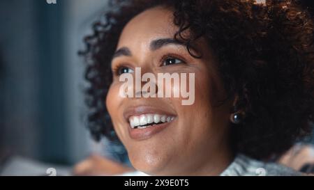 Beautiful Emotional Black Girl Sitting and Watching Television at Home. She is Laughing while Enjoying Funny Videos Online via Smart TV Set. Cozy and Stylish Room in Loft Apartment with Warm Light. Stock Photo