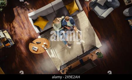 Top View Apartment: Happy Couple Using Laptop in the Stylish Living Room. Sitting on the Carpet and Smile. On the Floor: Girlfriend and Boyfriend use Computer for Online Shopping, e-business. Stock Photo