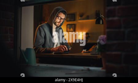 Professional Male Programmer Writing Code for New Project and Working Remotely on Laptop From Home Office. Freelance Software Engineer Designs App From his Apartment. View From Outdoors into Window. Stock Photo