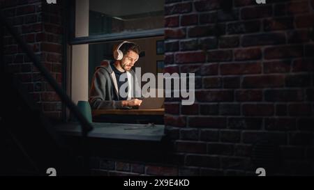 Software Developer Programming on a Laptop Late at Night. Male Specialist Browsing Internet, Listening to Podcast, Does Creative Marketing. Remote Work from Home. View From Outdoors into Window. Stock Photo