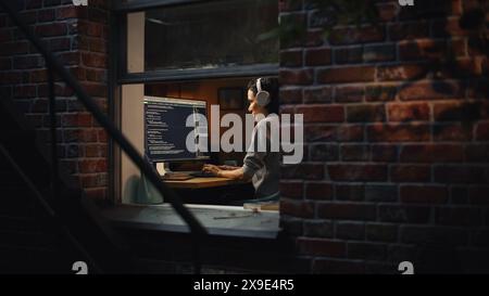 Latin Female Digital Startup Entrepreneur Working on Computer Late at Night. Software Developer Working on Innovative e-Commerce App. Overworking Concept. Shot Into the Window From Outside. Stock Photo