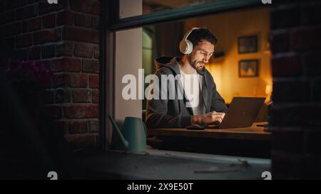Professional Software Engineer Working From Home Office. Tired IT Specialist Feeling Sleepy and Finising Important Project. Application Development. View From Outdoors into Window. Stock Photo