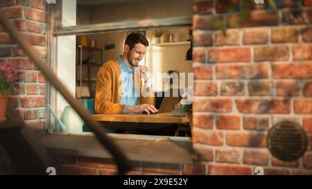 Handsome Creative Male Entrepreneur Working Remotely on Laptop Computer from Home. Freelance Businessman Using Portable Device, Developing e-Business Project. Into Window From Outdoors View. Stock Photo