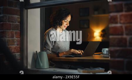 Smiling Hispanic Woman at Home Using Laptop During Warm Cozy Evening. Professional Freelancer Working Remotely From Stylish Home. Shot From Outside Into the Window of Apartment. Stock Photo