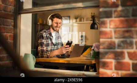 Handsome Caucasian Man Working on Laptop Computer while Sitting Behind His Desk at Home Office. Freelance Entrepreneur Listening to Music, Checking Smartphone. View From Outdoors into Window. Stock Photo